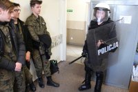 Uczniowie klasy mundurowej - jeden z nich przebrany jest w policyjny pancerz zwany PZ - ręku trzyma tarczę i pałkę, ma na sobie biały kask i ochraniacze na korpusie, rękach i nogach
