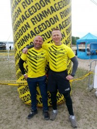 Dwaj policjanci w strojach sportowych i żółtych koszulkach z napisem Runmageddon, stoją obok siebie przed żółtym, walcowatym balonem z napisami Runmageddon.