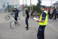 Na zdjęciu policjant z Wydziału Ruchu Drogowego przygląda się uczestnikowi egzaminu na kartę rowerową, który zakłada kask. W/w stoją na szkolnym boisku, przyglądają im się uczniowie.