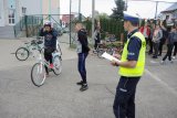 Na zdjęciu policjant z Wydziału Ruchu Drogowego przygląda się uczestnikowi egzaminu na kartę rowerową, który zakłada kask. W/w stoją na szkolnym boisku, przyglądają im się uczniowie.