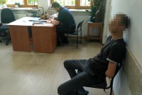 Na zdjęciu widać policjanta który wypełnia dokumentację przy biurku, obok niego, na krześle siedzi zatrzymany mężczyzna.