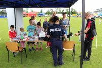 Na zdjęciu widać policjantkę przy stoisku ,która prezentuje dzieciom wyposażenie policyjne, które zakłada jedno z dzieci.