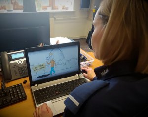 policjantka siedząca przed monitorem laptopa. na monitorze animacja , idący mężczyzna patrzy na telefon przed nim otwór z napisem uwaga ! uzależnienie