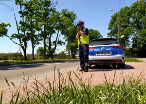 policjant kontrolujący prędkość za obok nie radiowóz za nim widok na drogę na poboczu drzewa