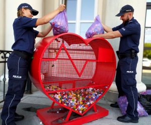 policjantka i policjant wrzucają nakrętki znajdujące się w workach do metalowego pojemnika w kształcie serca