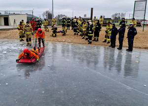 strażak leżący na saneczkach ratunkowych na tafli lodu na drugim planie policjanci i strażacy