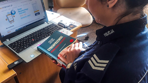 policjantka siedząca przed laptopem w ręku trzyma książkę zatytułowana cyberprzemoc wśród dzieci i młodzieży
