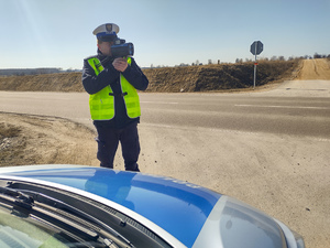 Policjant stojący przy drodze, mierzący urządzeniem prędkość samochodów.