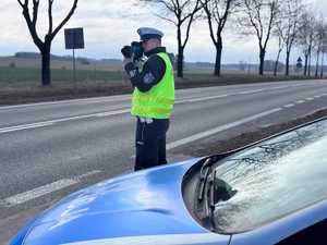 Policjant w białej czapce mierzy prędkość pojazdów obok radiowóz