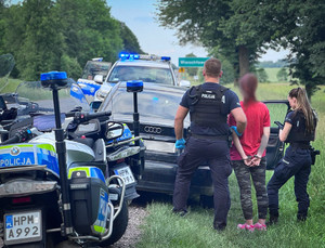 Kobieta w kajdankach trzymana przez policjanta, obok radiowóz i policjantka